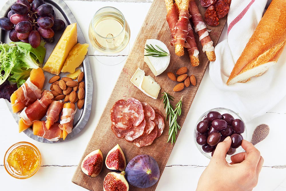 Charcuterie board set up with cheese, meats, fruit, olives, bread and wine. Hand image grabbing an olive.