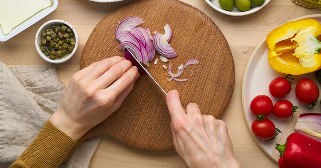 Person cutting a red onion to assemble a greek salad for a vegan hummus grain bowl for lunch.